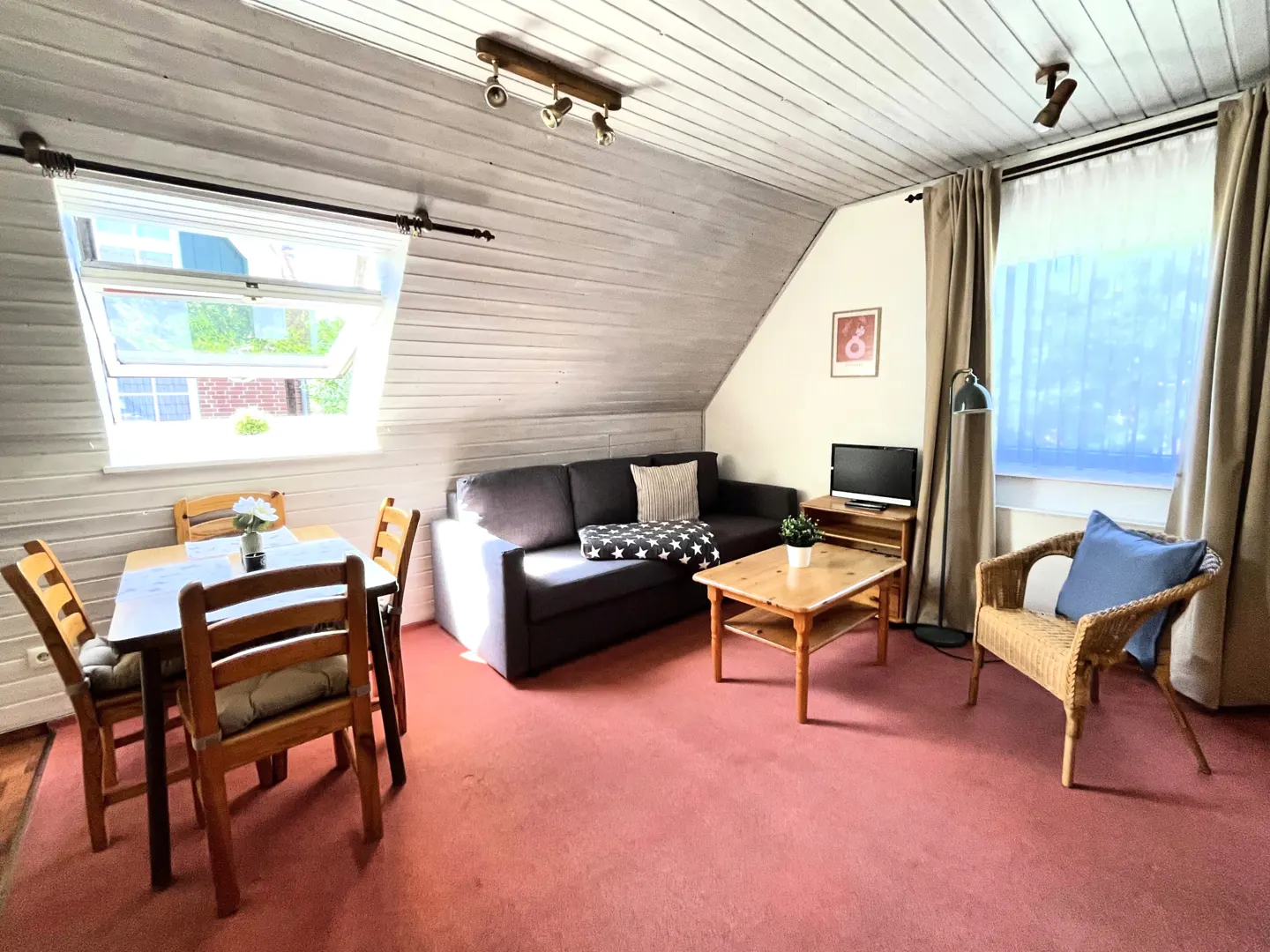 Attic living room with white wood paneled walls, pink carpet, gray sofa, wood table and chairs, and a window with green foliage outside.