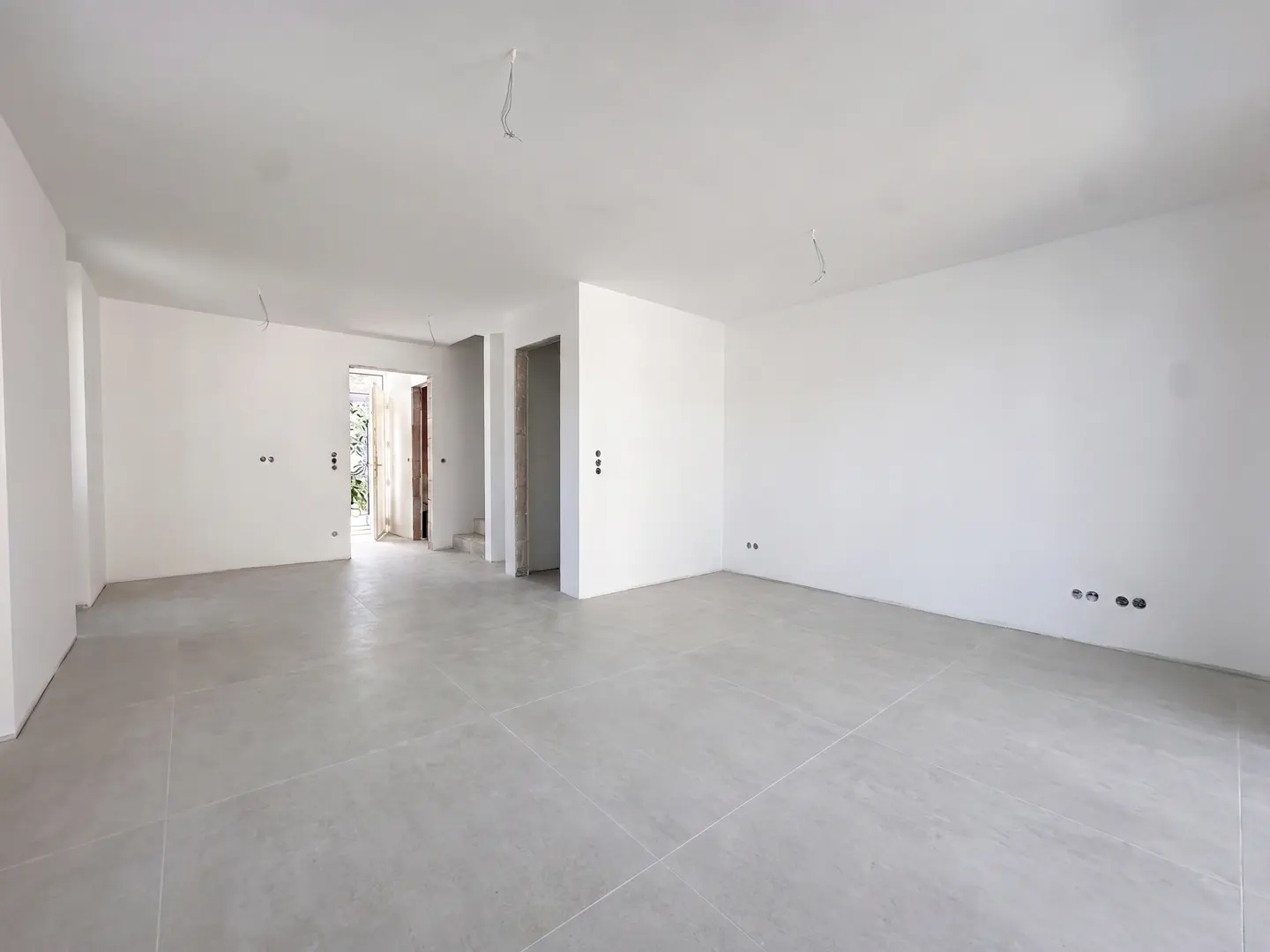 Bright, empty room with white walls and gray tile floor. Doorways lead to other rooms and the outside. Electrical wiring visible.