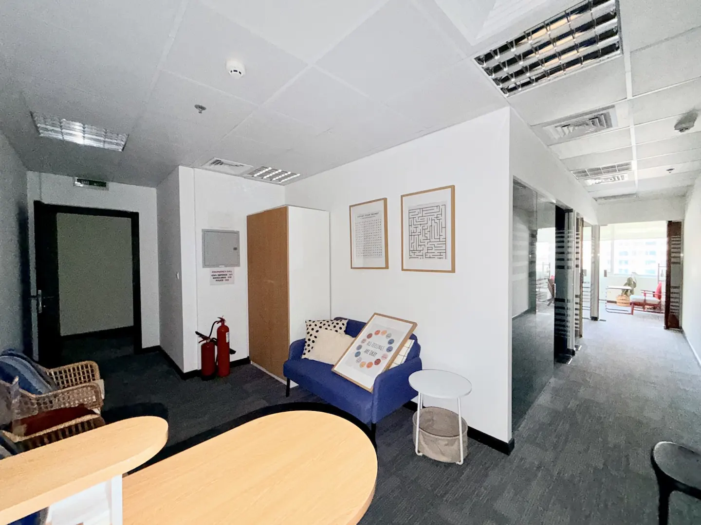 Modern office waiting area with a blue sofa, wooden cabinet, fire extinguisher, and a view into a bright hallway.