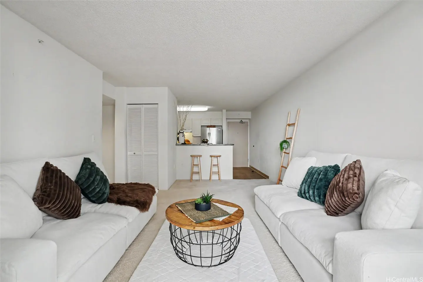 Bright living room with white sofas, brown and green pillows, and a round wood and metal coffee table on a white rug. Kitchen in background.