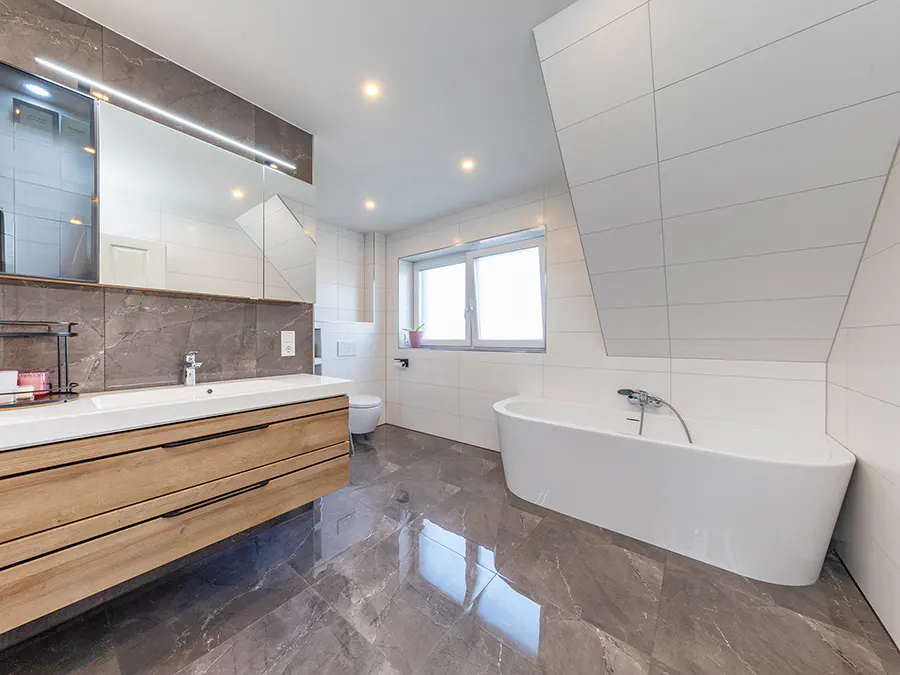 Bright bathroom with gray marble floors, white walls, and a large mirror above a wooden vanity. A white tub sits near a window.