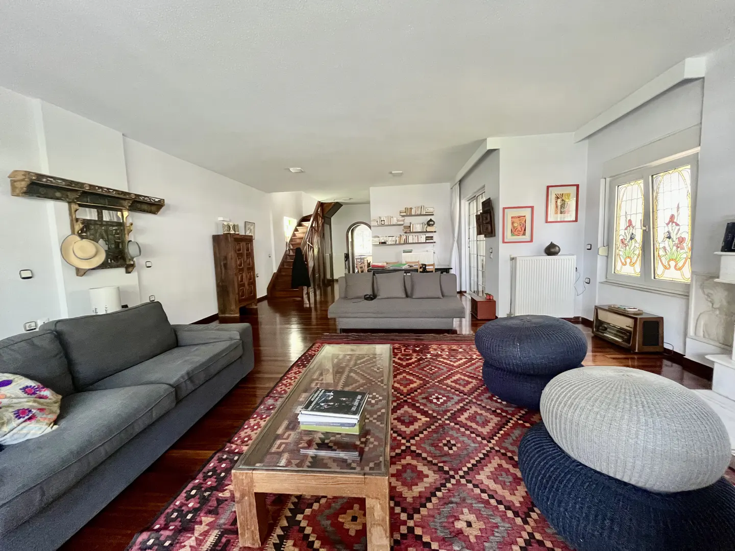Bright living room with hardwood floors, gray sofas, and a red patterned rug. A glass coffee table sits in the center, with blue and gray poufs nearby. Stairs are visible in the background.