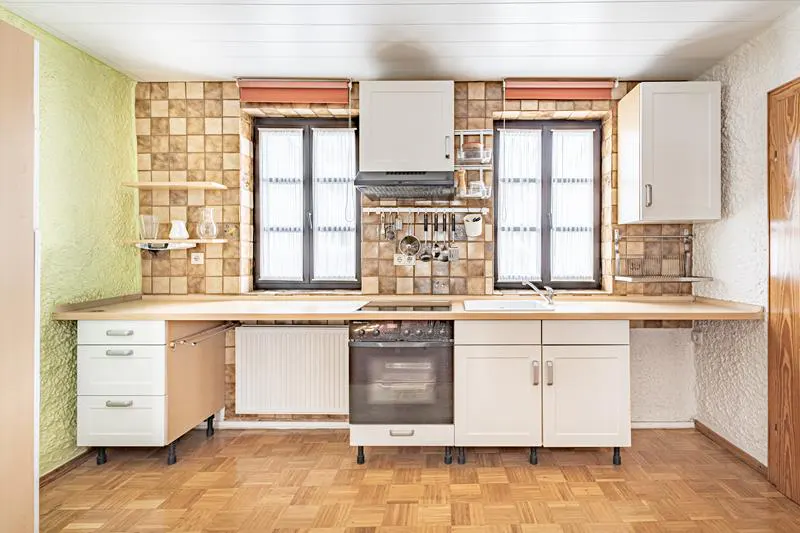 A kitchen with white cabinets, a wood countertop, and brown tiled walls. Two windows with white curtains are above the sink and stove.
