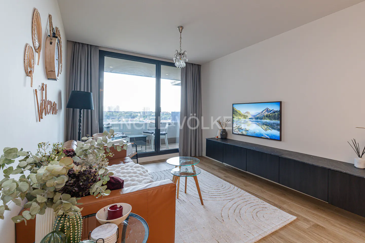 Modern living room with a tan leather sofa, glass coffee table, wall-mounted TV, and a balcony view through large windows.
