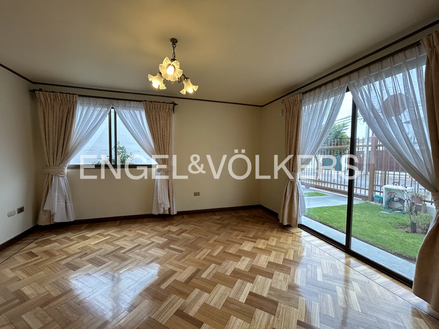 A bright, empty room with parquet floors, beige walls, and large windows with sheer curtains. A floral chandelier hangs from the ceiling.