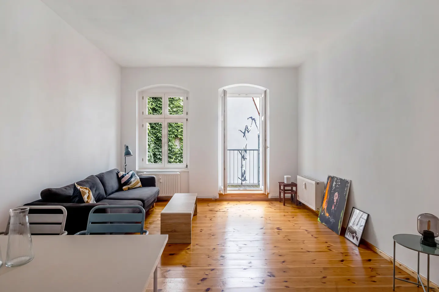 Bright living room with wood floors, white walls, and a gray sofa. A window with ivy and a balcony door let in natural light.