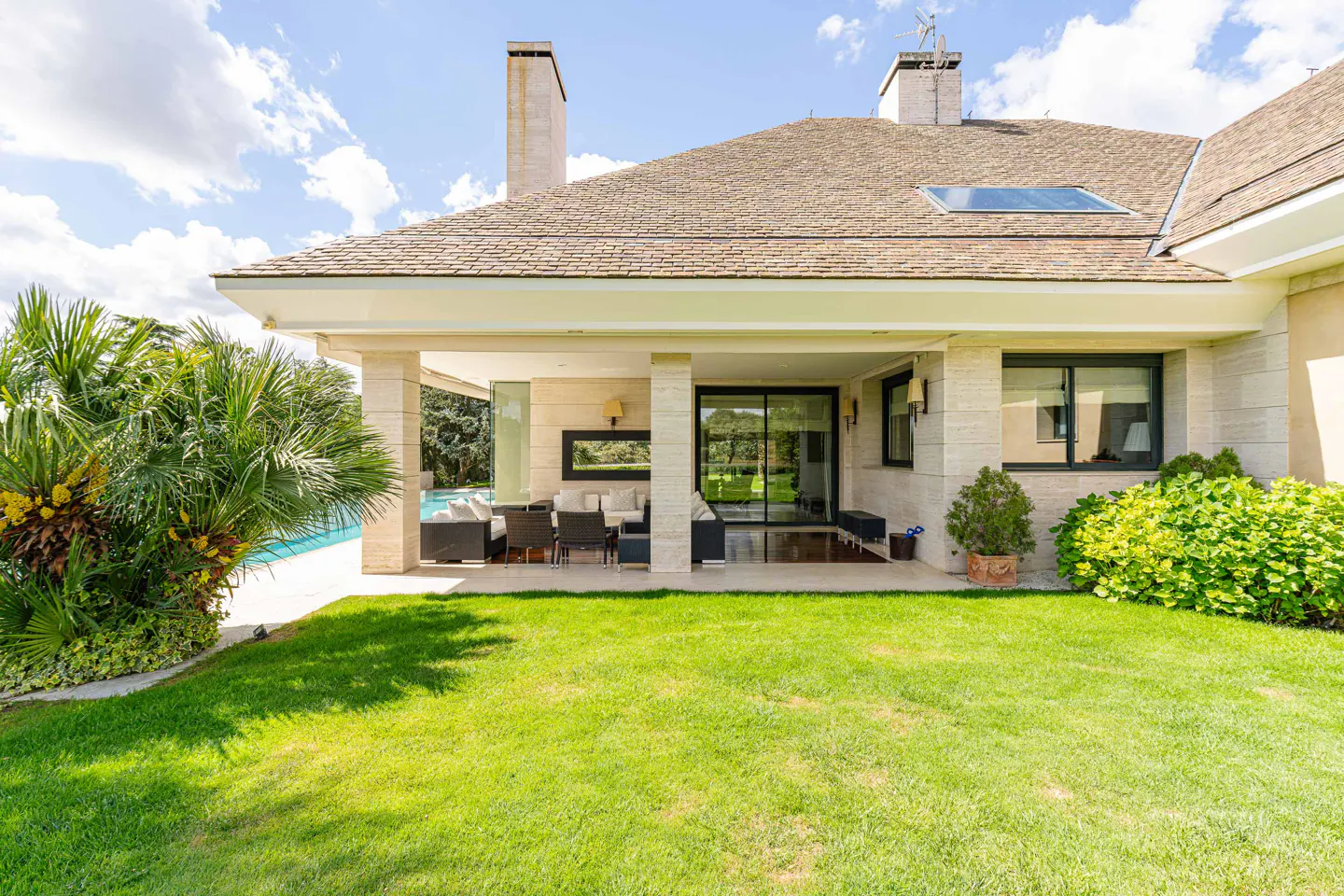 Exterior view of a beige stone house with a covered patio, green lawn, and a glimpse of a blue swimming pool.