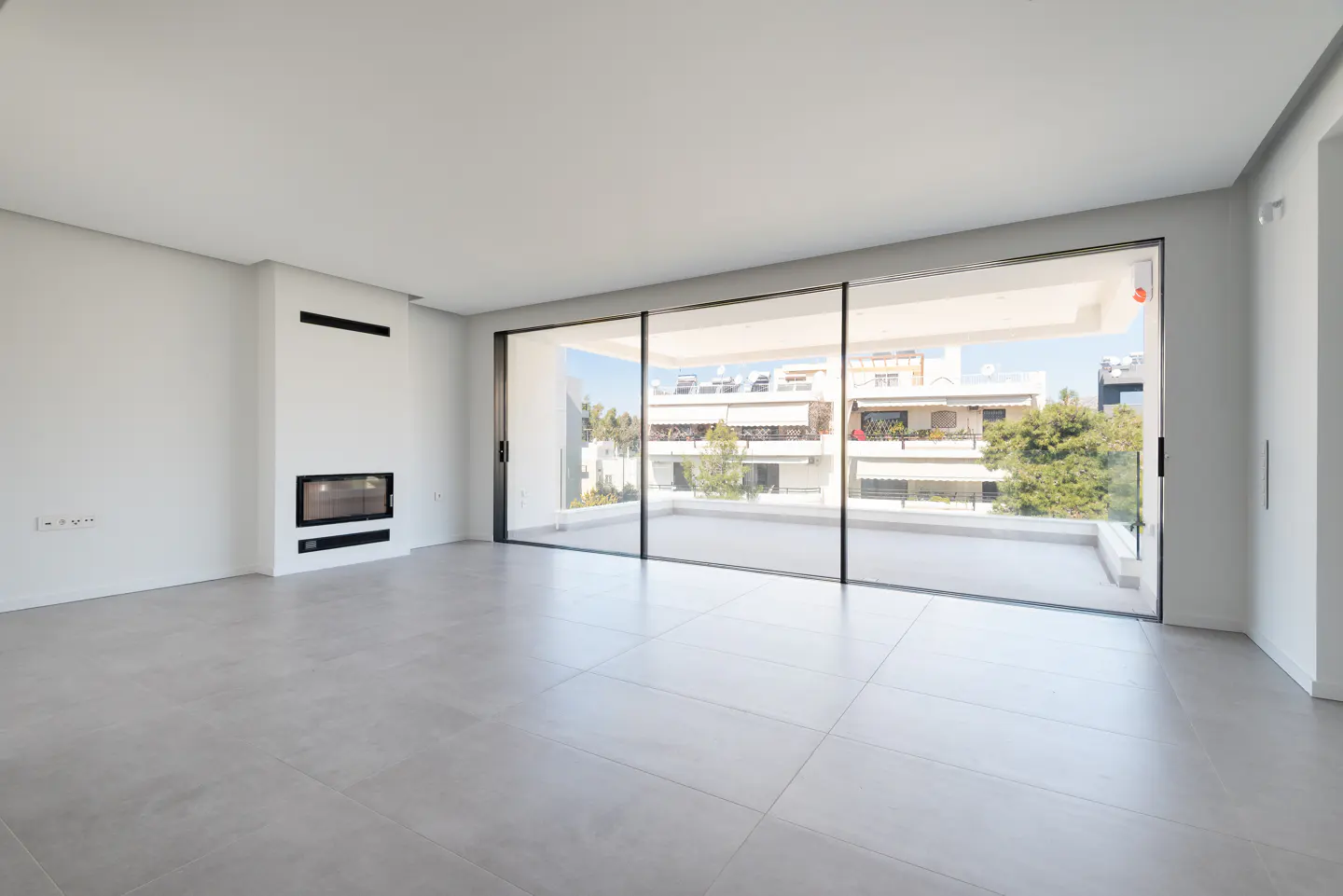 Bright, empty room with gray tile floor, white walls, and a modern fireplace. Large sliding glass doors lead to a balcony with city views.