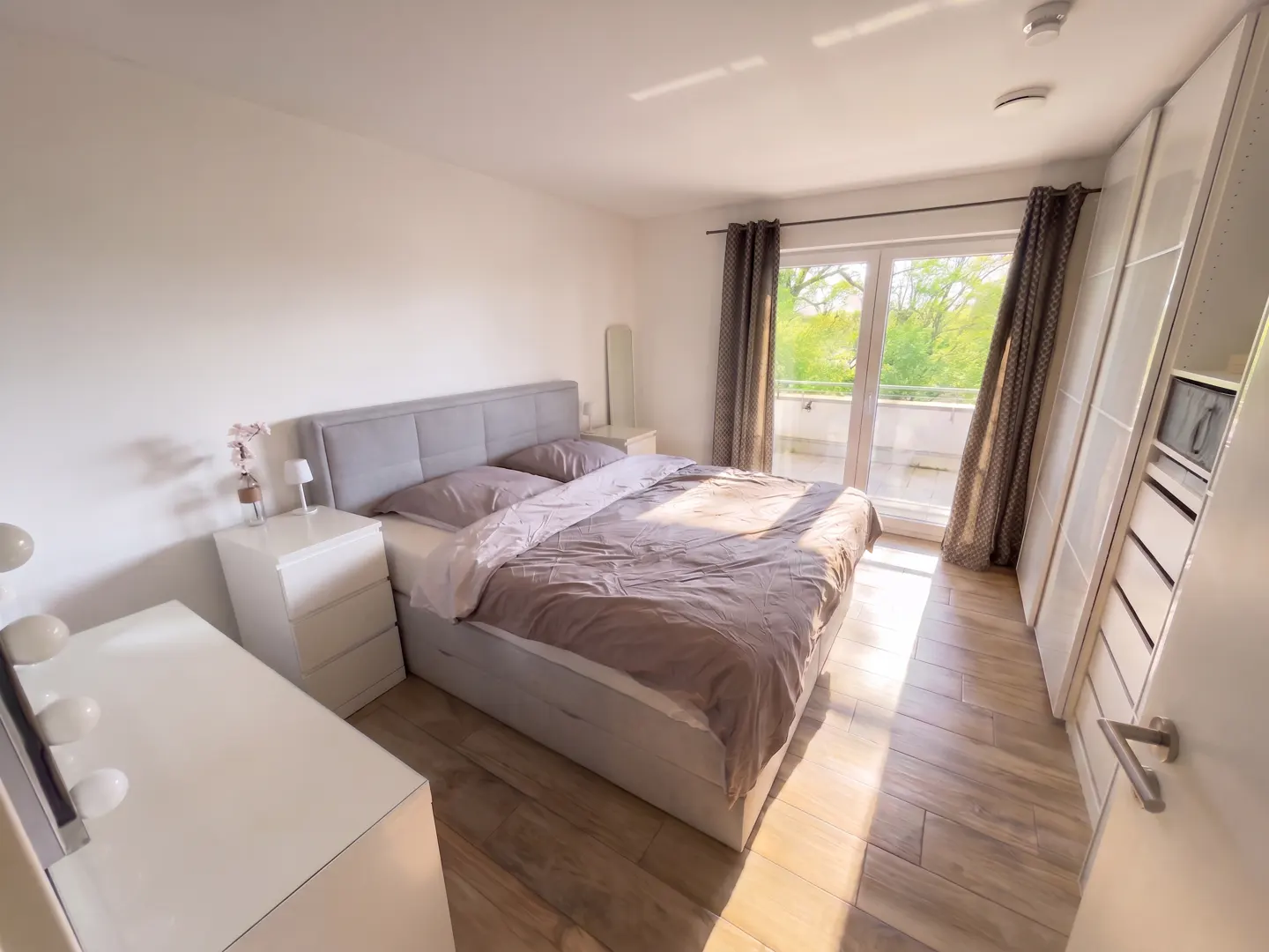 Bright bedroom with a gray bed, white nightstands, and a balcony view. Wood floors and white walls create a clean, modern aesthetic.