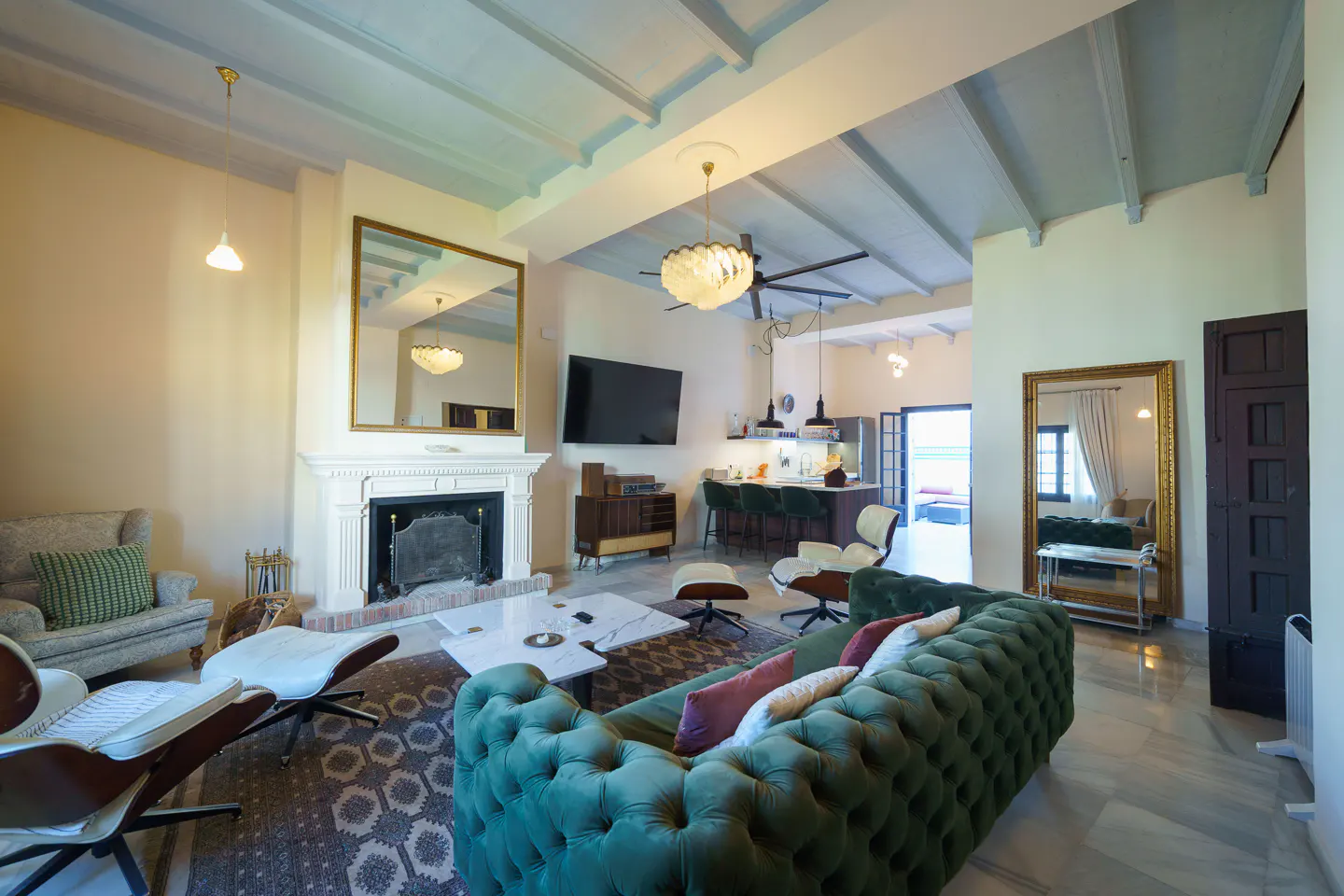 Bright living room with a green tufted sofa, fireplace, and a large mirror. The room opens to a kitchen with bar stools.