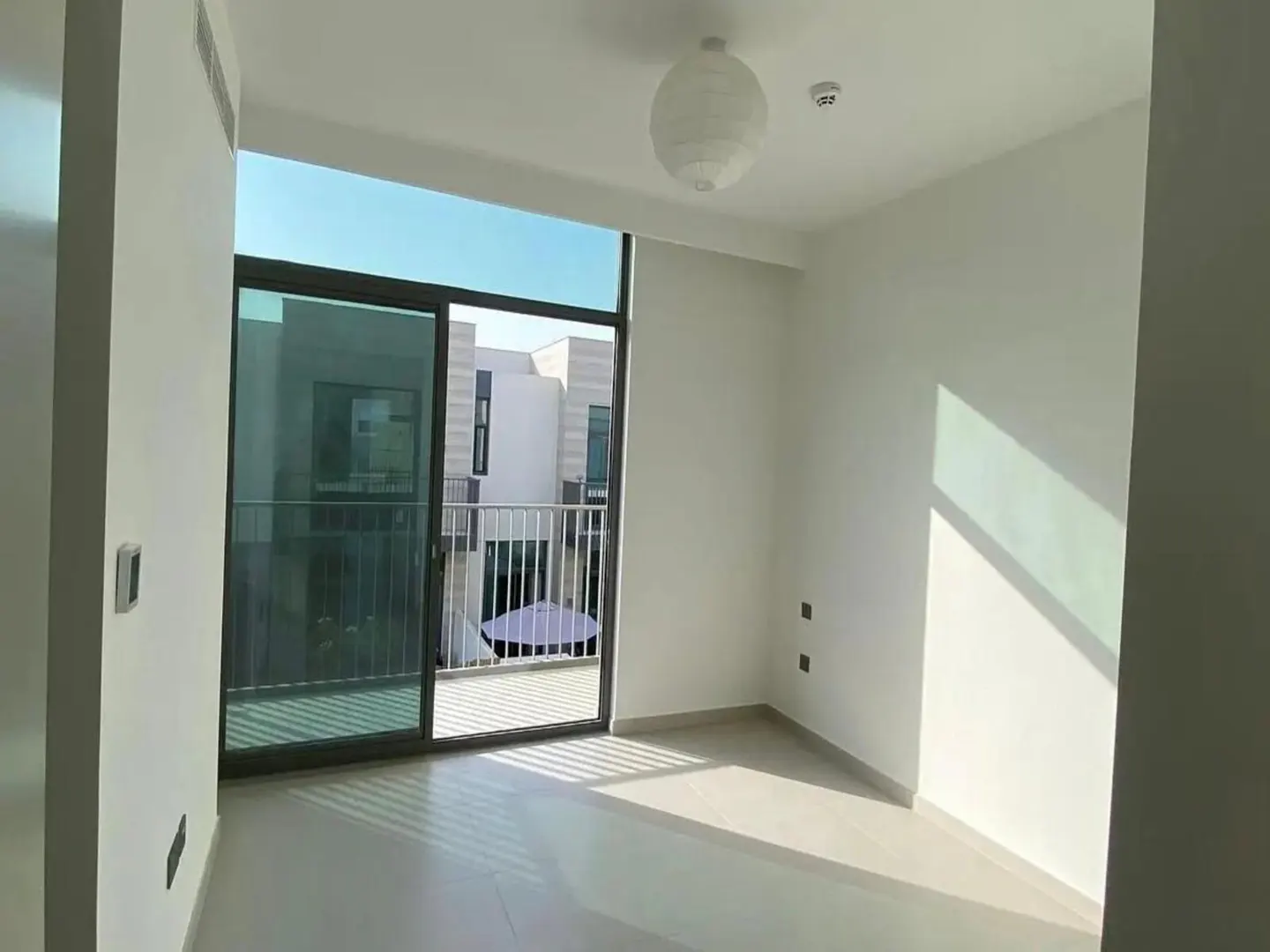 Empty minimalist room with white walls, tile floors, a paper globe pendant light, and a balcony view of modern buildings.