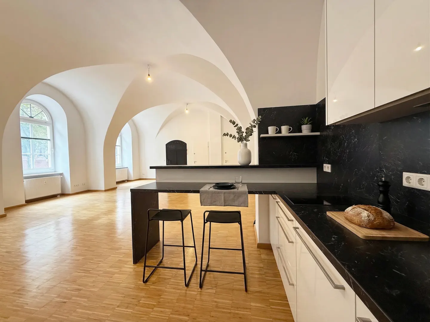 A modern kitchen with black countertops, white cabinets, and two black stools at the island. The room has arched ceilings and wood floors.