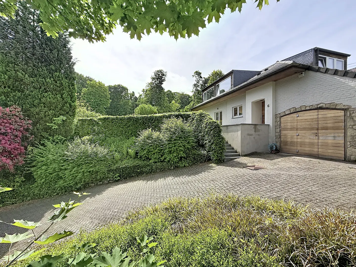 Exterior view of a white house with a wooden garage door and a brick driveway surrounded by lush greenery.