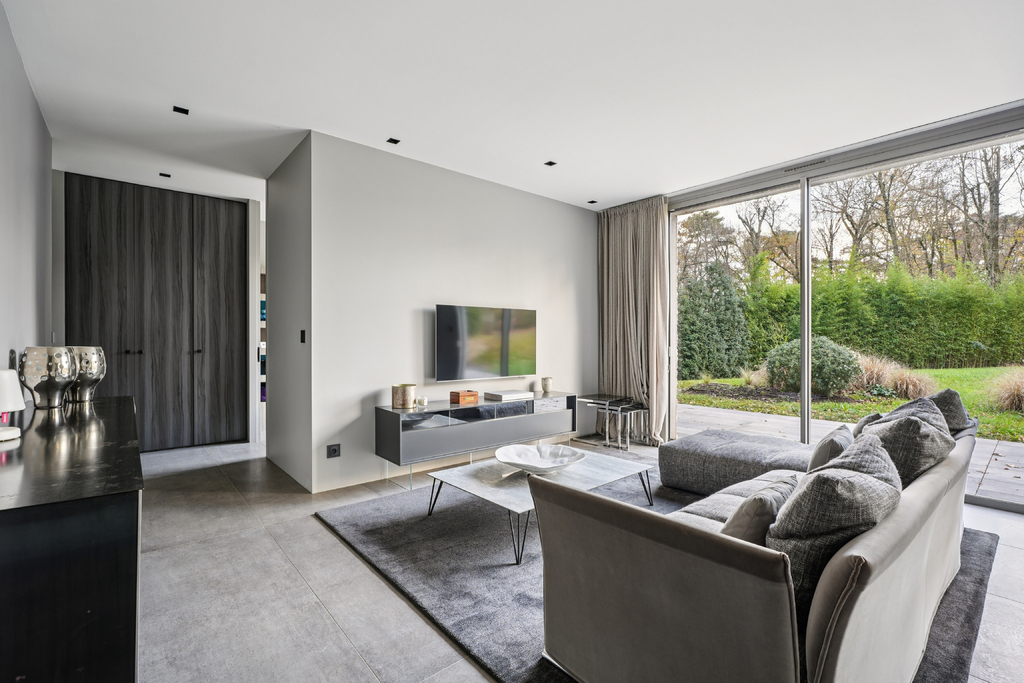 A modern living room with gray walls, a gray sofa, a TV, and a large window overlooking a green yard.