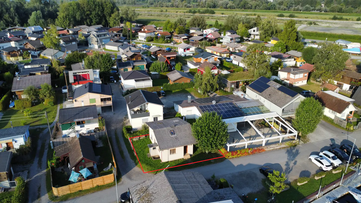 Aerial view of a small, light-colored house with a gray roof, outlined in red, in a neighborhood with trees and other houses.