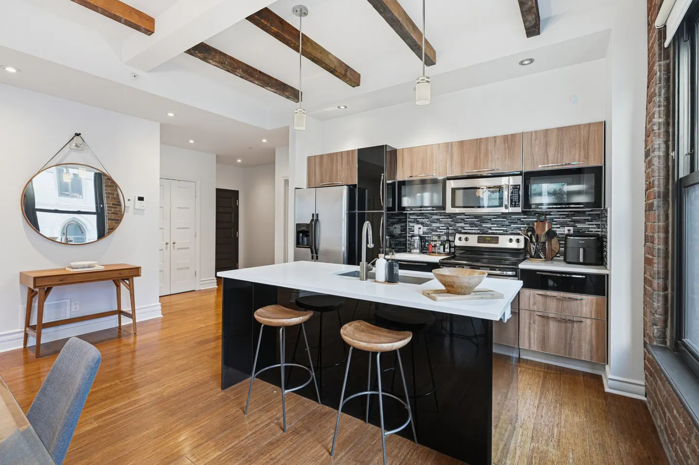 Bright, modern kitchen with wood floors, black island with white countertop, and wooden stools. Exposed wood beams on the ceiling.