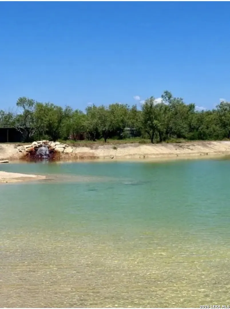 A scenic view of a turquoise lake with a small waterfall, surrounded by trees under a clear blue sky.