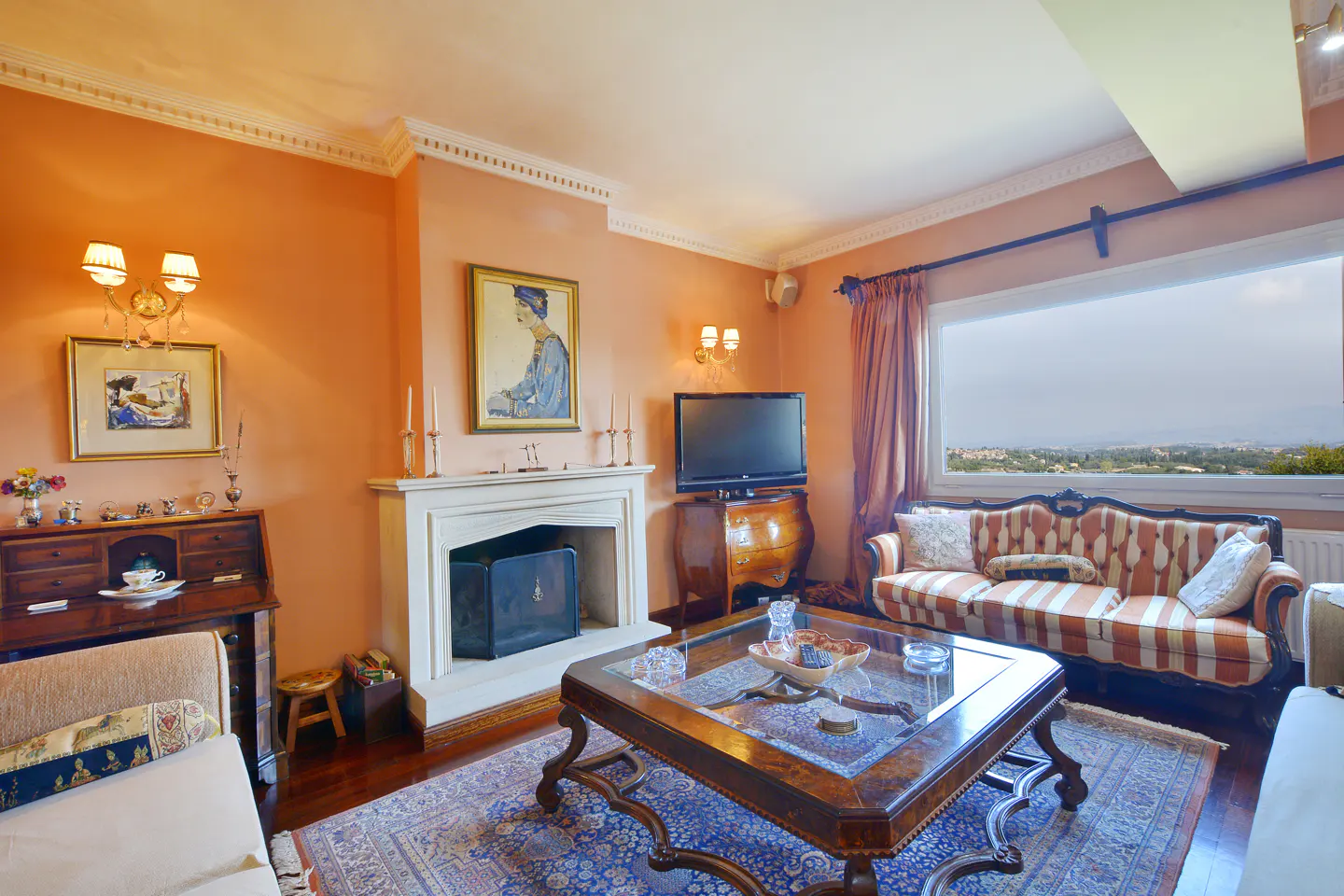 Living room with orange walls, fireplace, striped sofa, and a large window with a view. A blue patterned rug sits under a glass-topped coffee table.