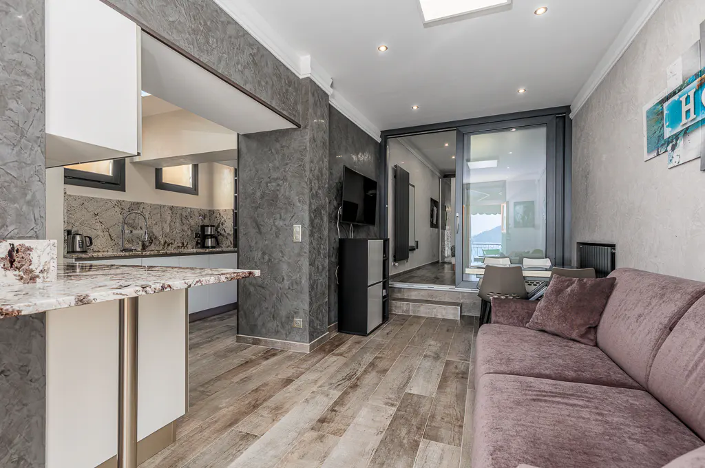 A modern apartment interior with a kitchen, living room, and balcony. The living room has a purple sofa and a sliding glass door to the balcony.