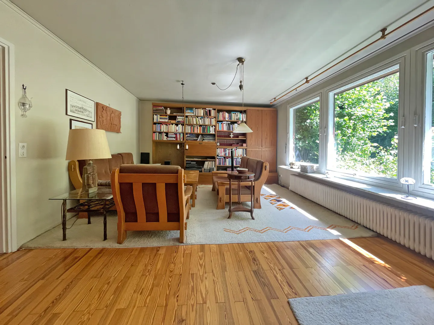 Living room with wooden floors, a large bookshelf, brown seating, a patterned rug, and large windows overlooking greenery.