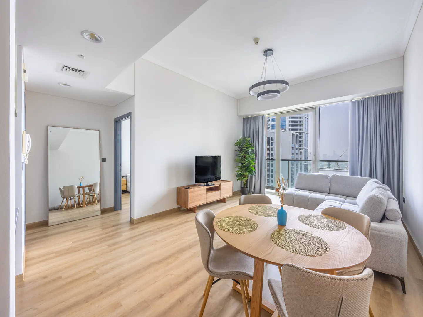 Bright apartment interior with wood floors, a round table with four chairs, and a gray sectional sofa near a window with city views.