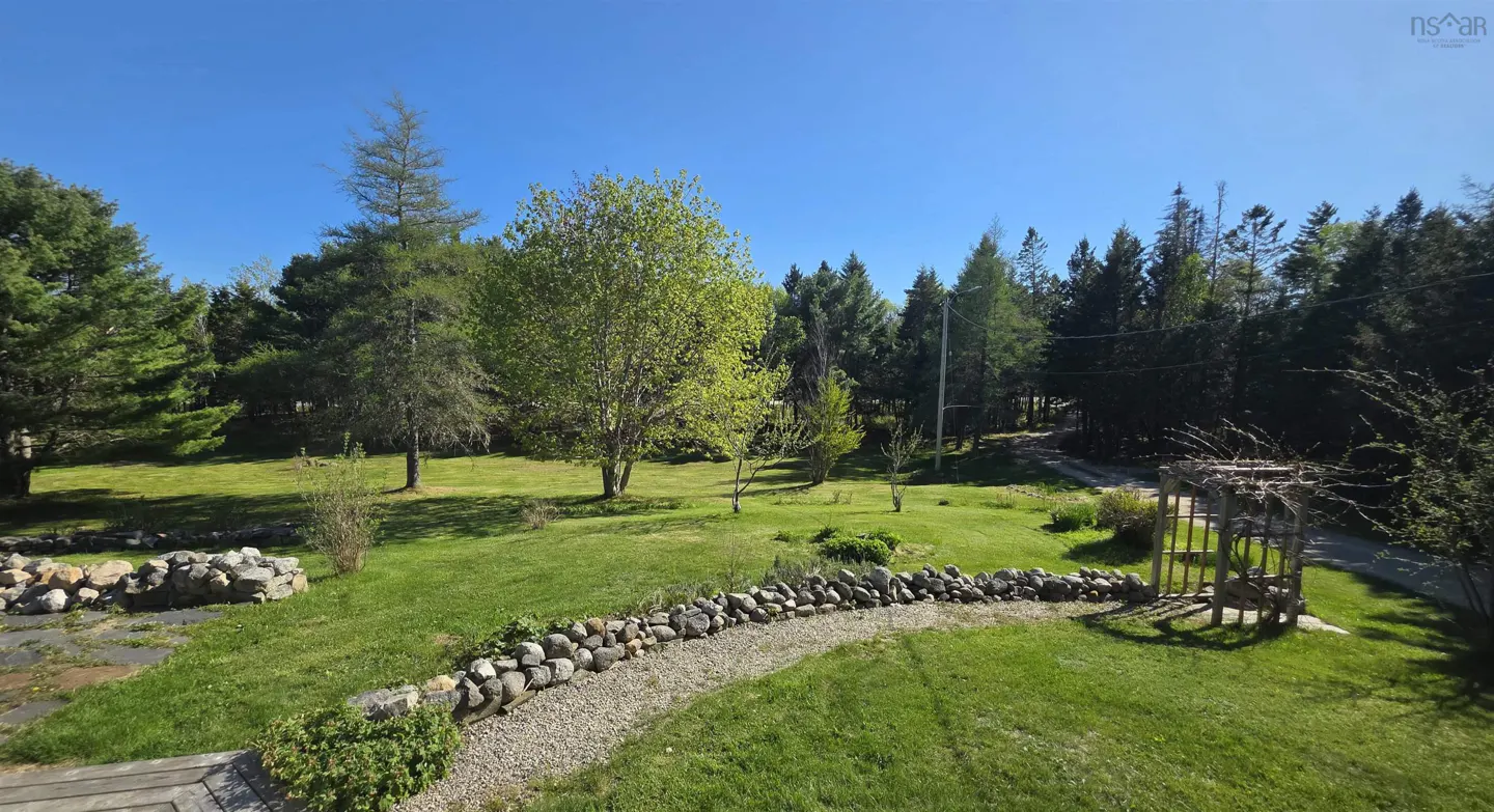 A lush green backyard with a stone retaining wall, a gravel path, a wooden trellis, and mature trees under a clear blue sky.