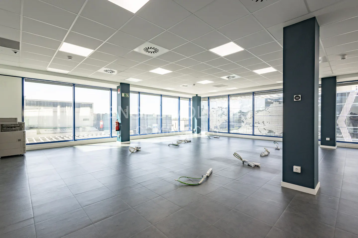 Empty modern office space with grey tiled floors, large windows, and dark blue structural columns.