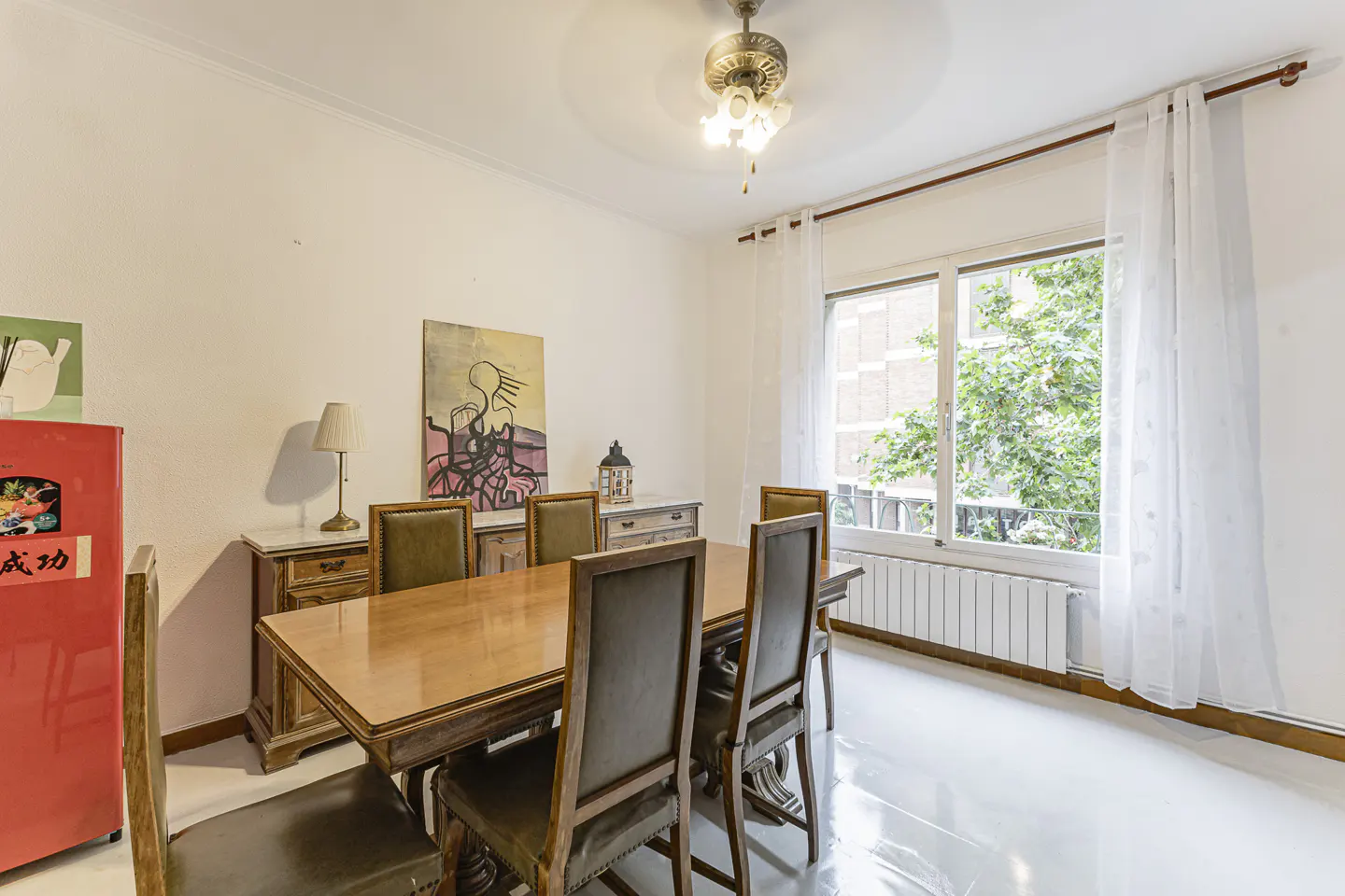 Bright dining room with a wooden table, chairs, and a red refrigerator. A window with white curtains lets in natural light.