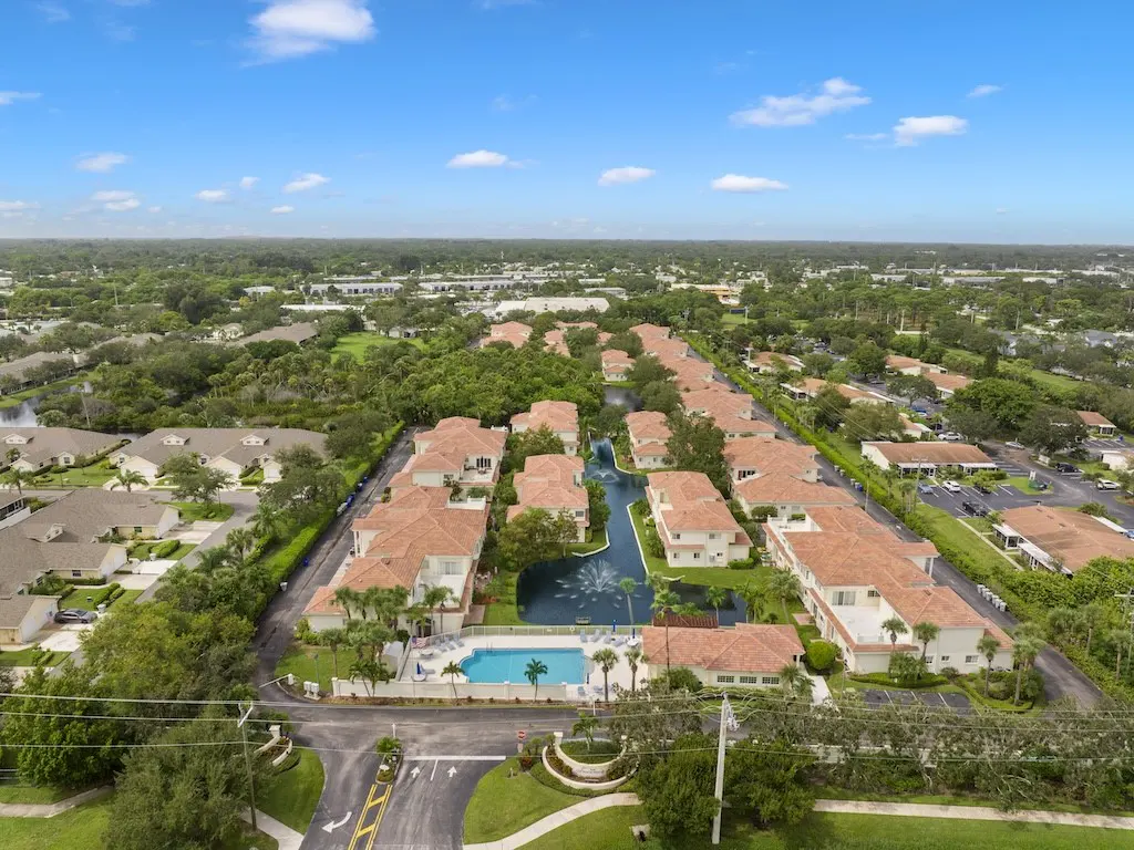 Aerial view of a residential community with red-tiled roofs, a pool, and a waterway with fountains, surrounded by lush greenery.