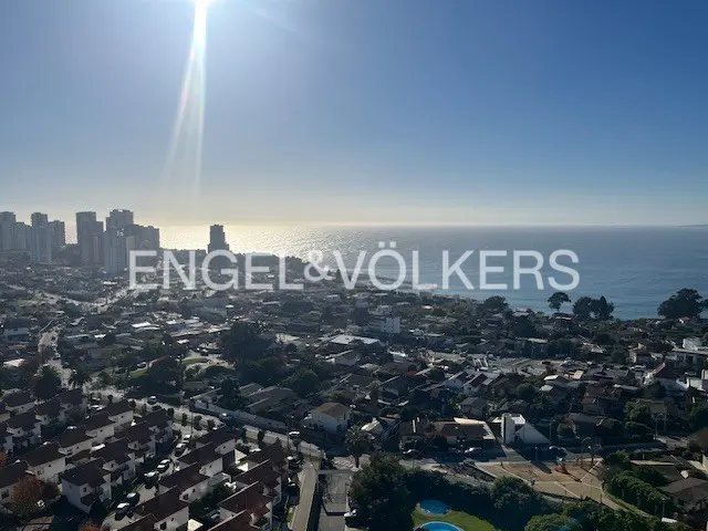 Real estate view of a city with buildings, houses, and the ocean under a bright blue sky. Engel & Völkers logo is overlaid.