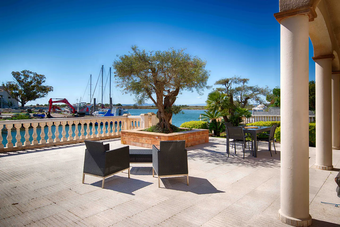 Outdoor patio with black chairs, a table, and a tree in a brick planter overlooking a blue harbor with boats under a clear blue sky.