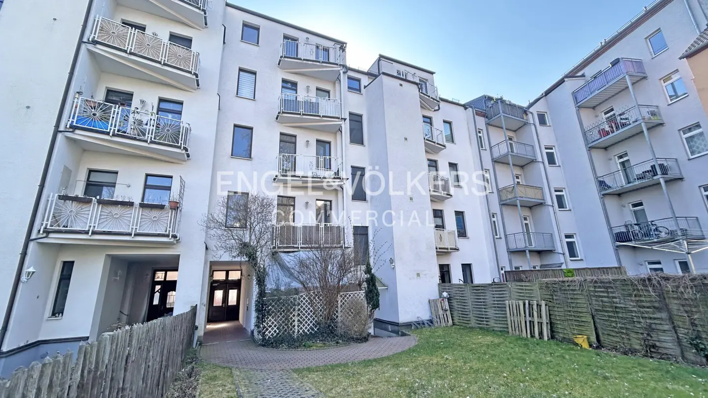 Exterior view of a white apartment building with balconies, a green lawn, and a wooden fence.