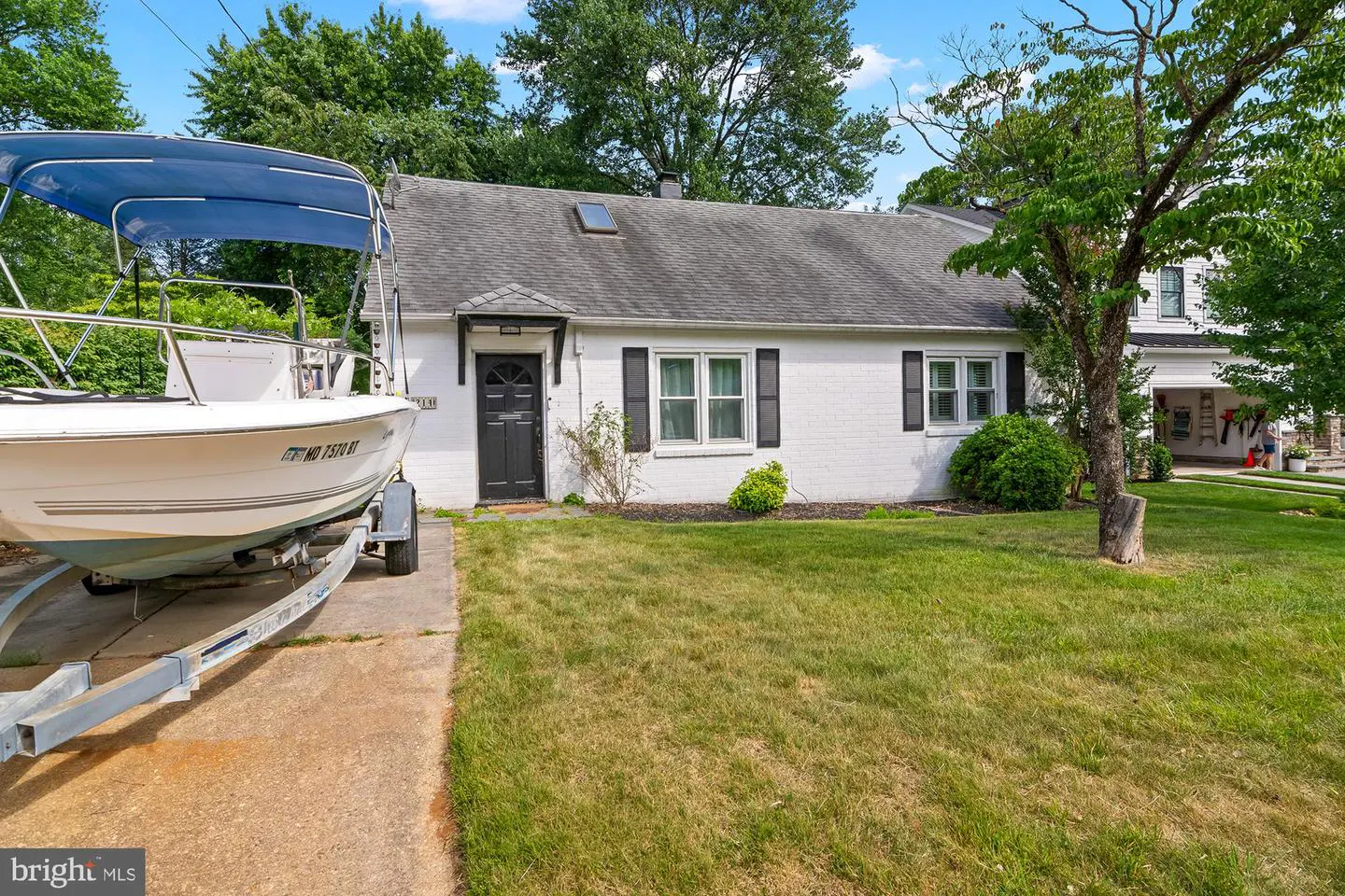 White brick cottage with black shutters and a boat on a trailer parked in the driveway on a sunny day.