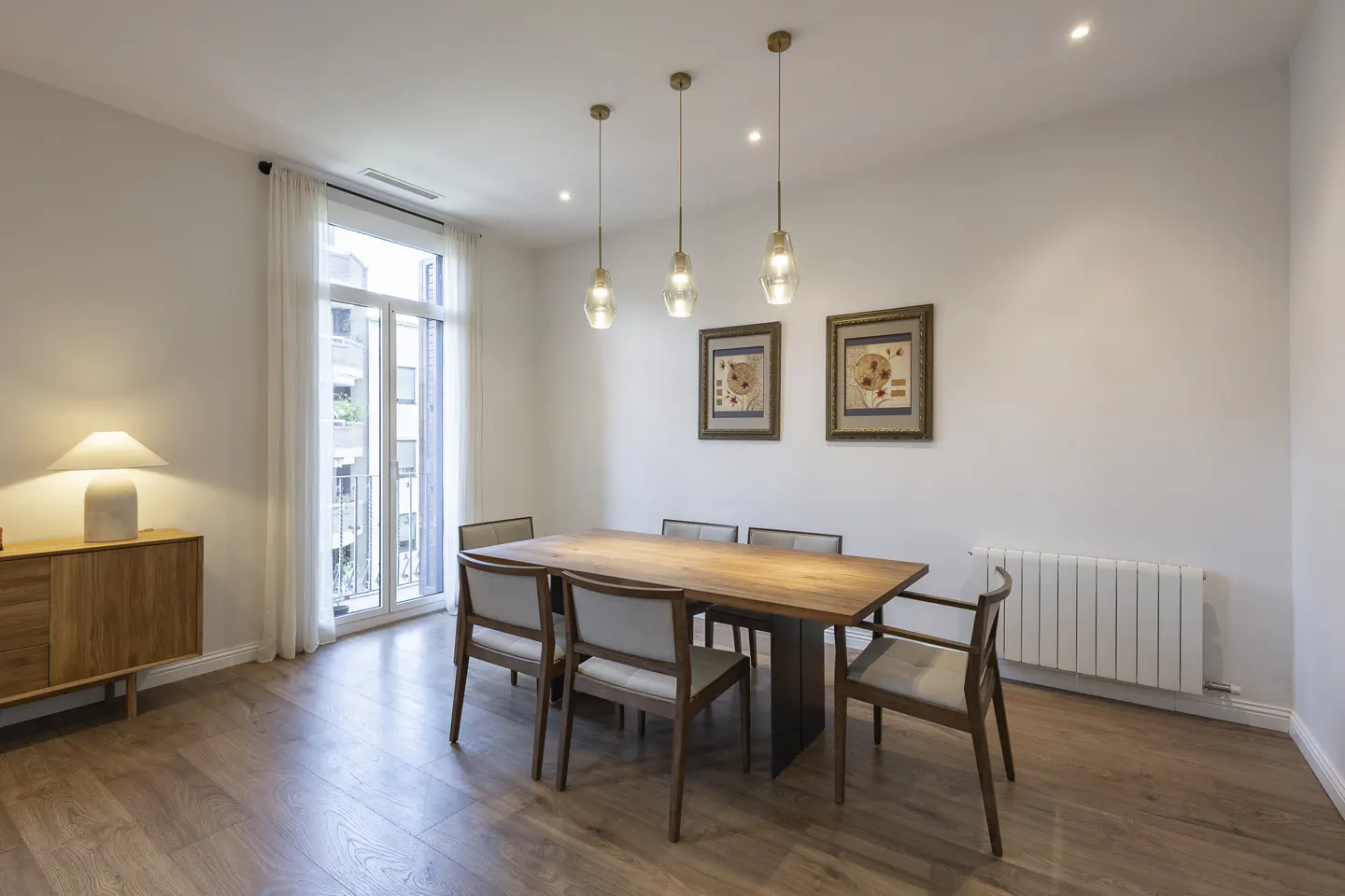 Bright dining room with wood floors, table, and six chairs. Three pendant lights hang above the table. A sideboard and lamp are on the left.