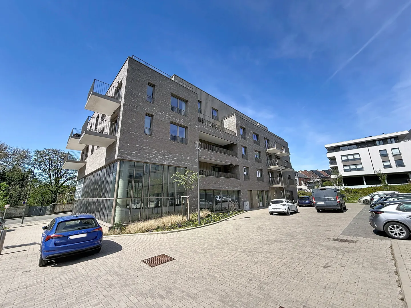 Modern gray brick apartment building with balconies and a glass-walled ground floor, surrounded by a parking lot with cars.