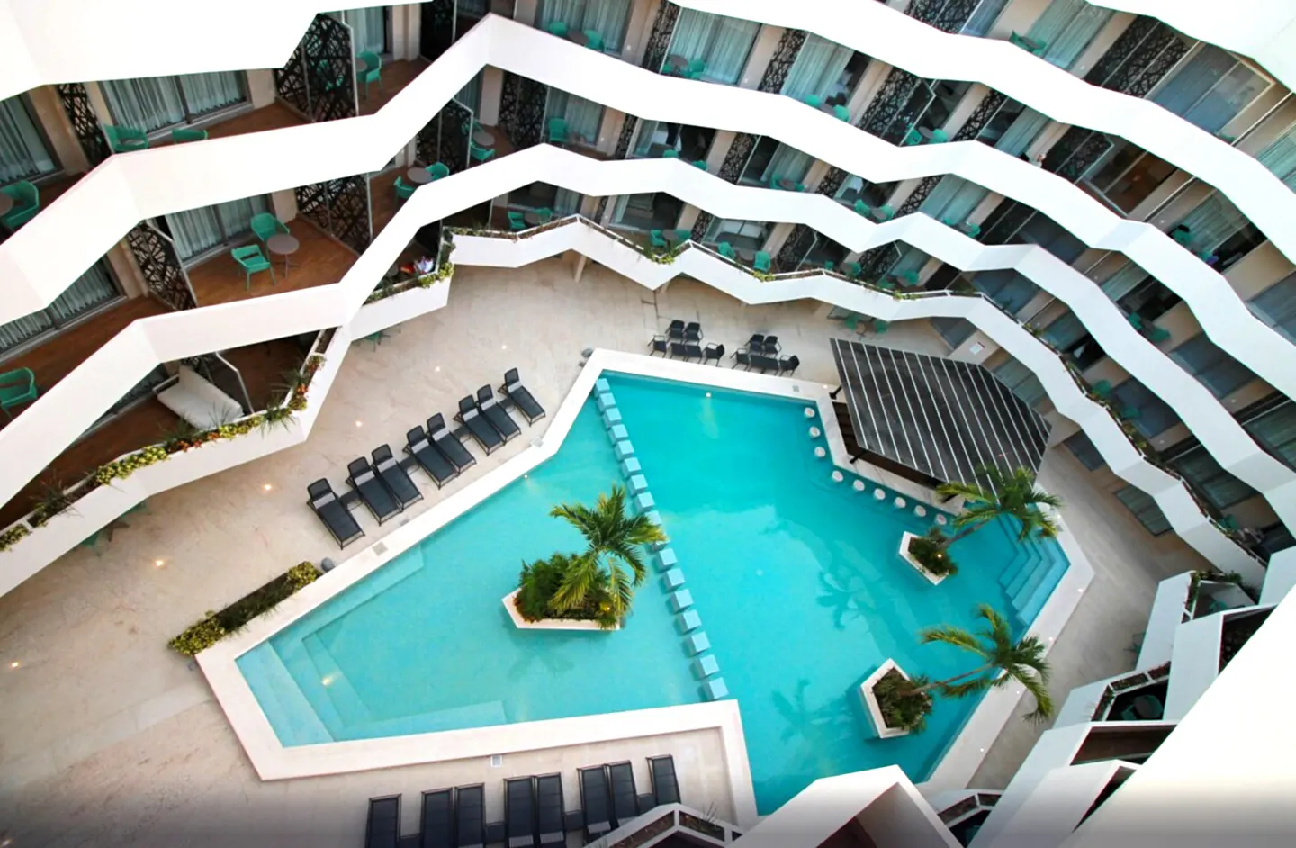 Aerial view of a hotel courtyard with a turquoise pool, palm trees, lounge chairs, and balconies with green furniture.