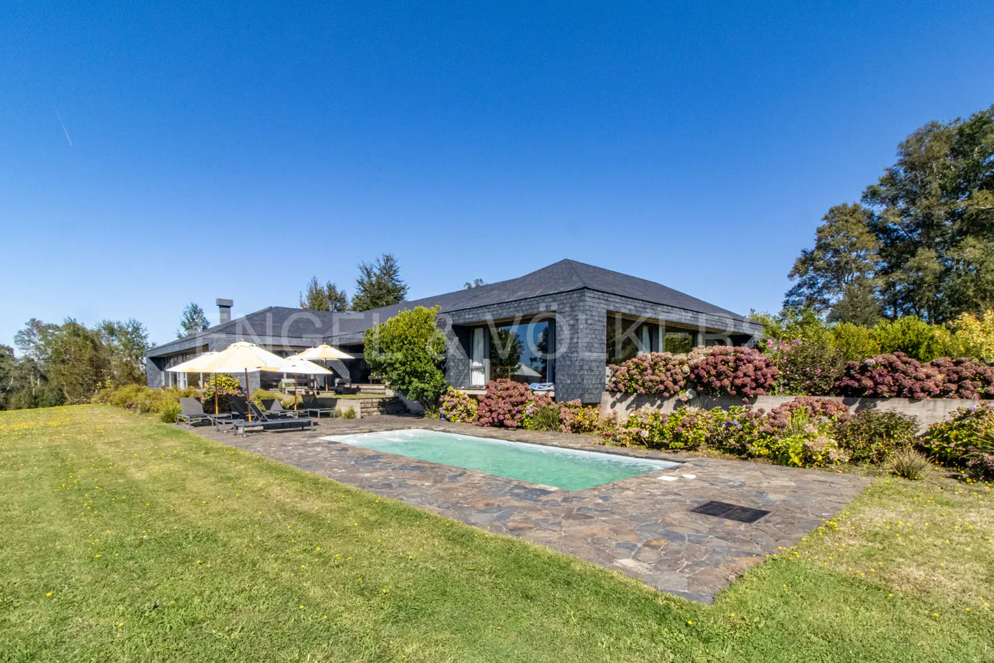 Exterior view of a modern, dark gray house with a pool, patio furniture, and lush green lawn under a clear blue sky.