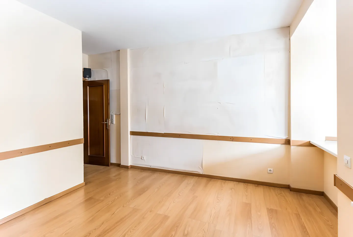 Empty room with light wood floors, cream walls, and a brown door. A window is on the right.