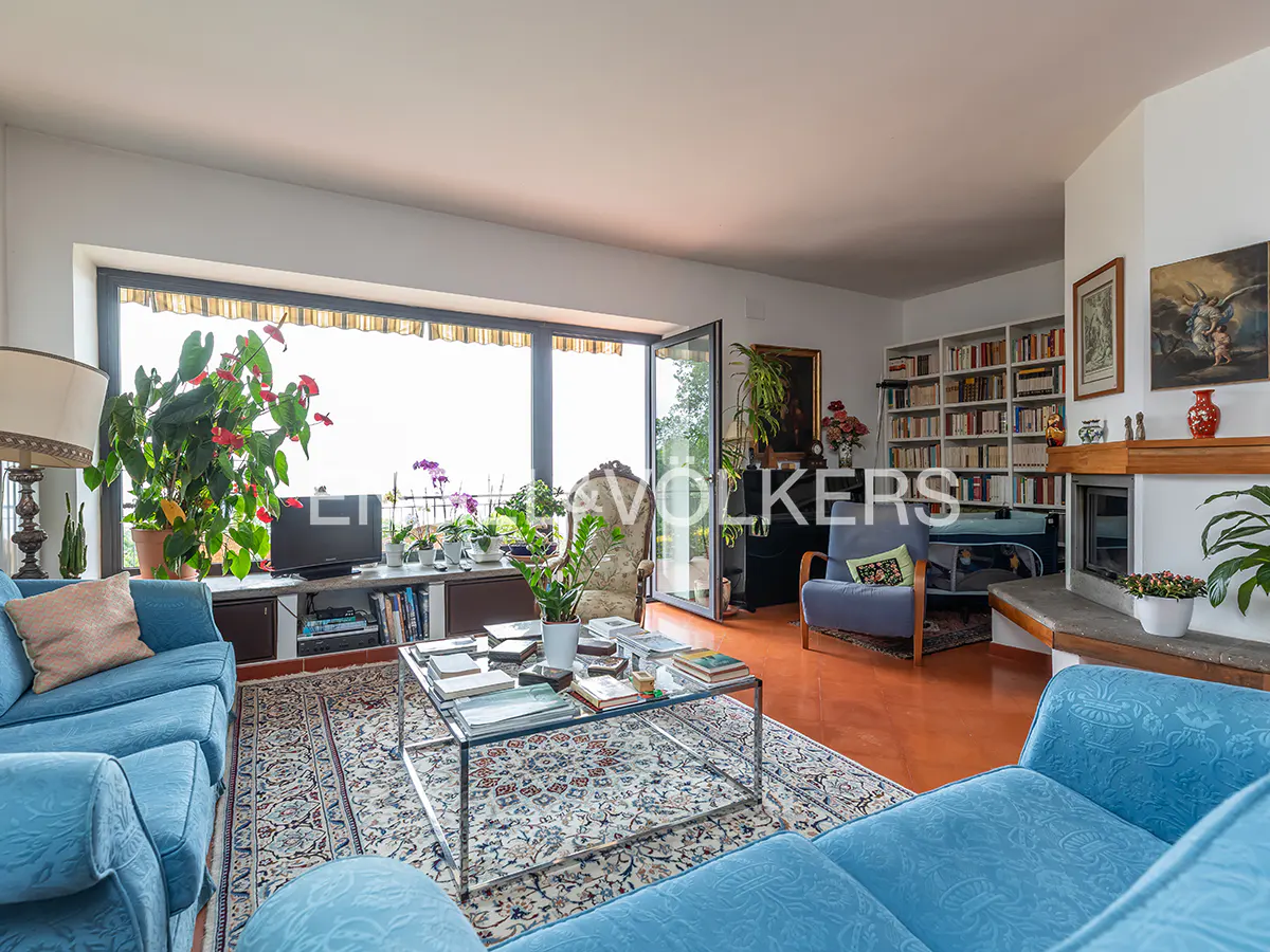 Bright living room with blue sofas, a glass coffee table, a large window, a fireplace, and a wall-to-wall bookshelf.