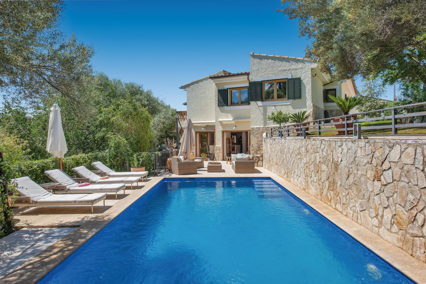A rectangular blue pool is surrounded by lounge chairs and a stone wall, with a cream-colored house in the background.