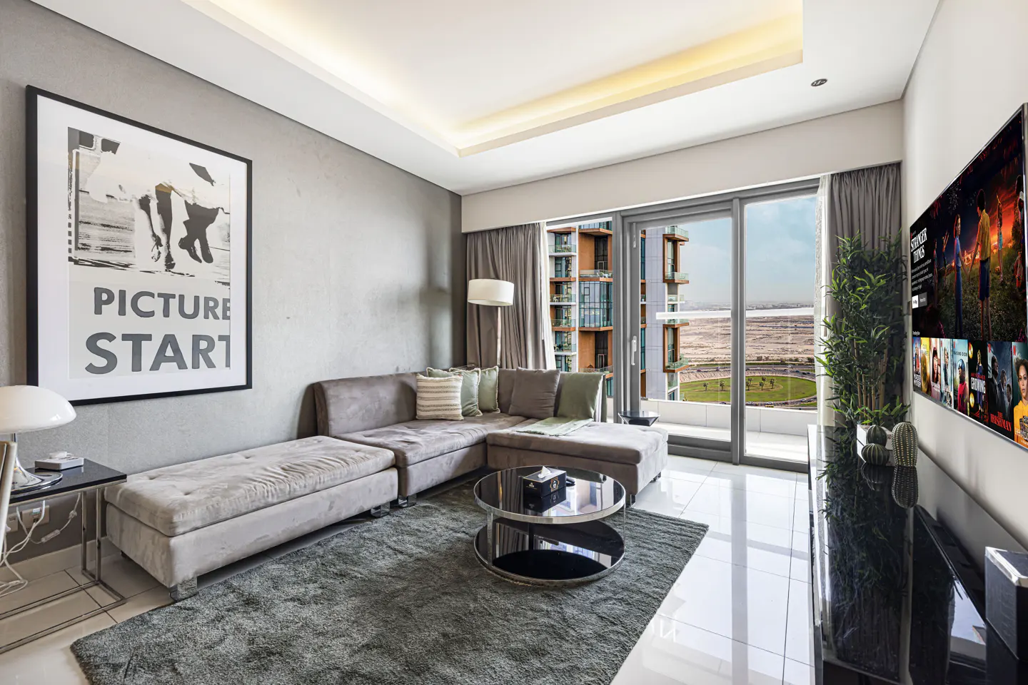 Living room with gray sectional sofa, round coffee table, and large framed art. Balcony with city view. TV on wall.
