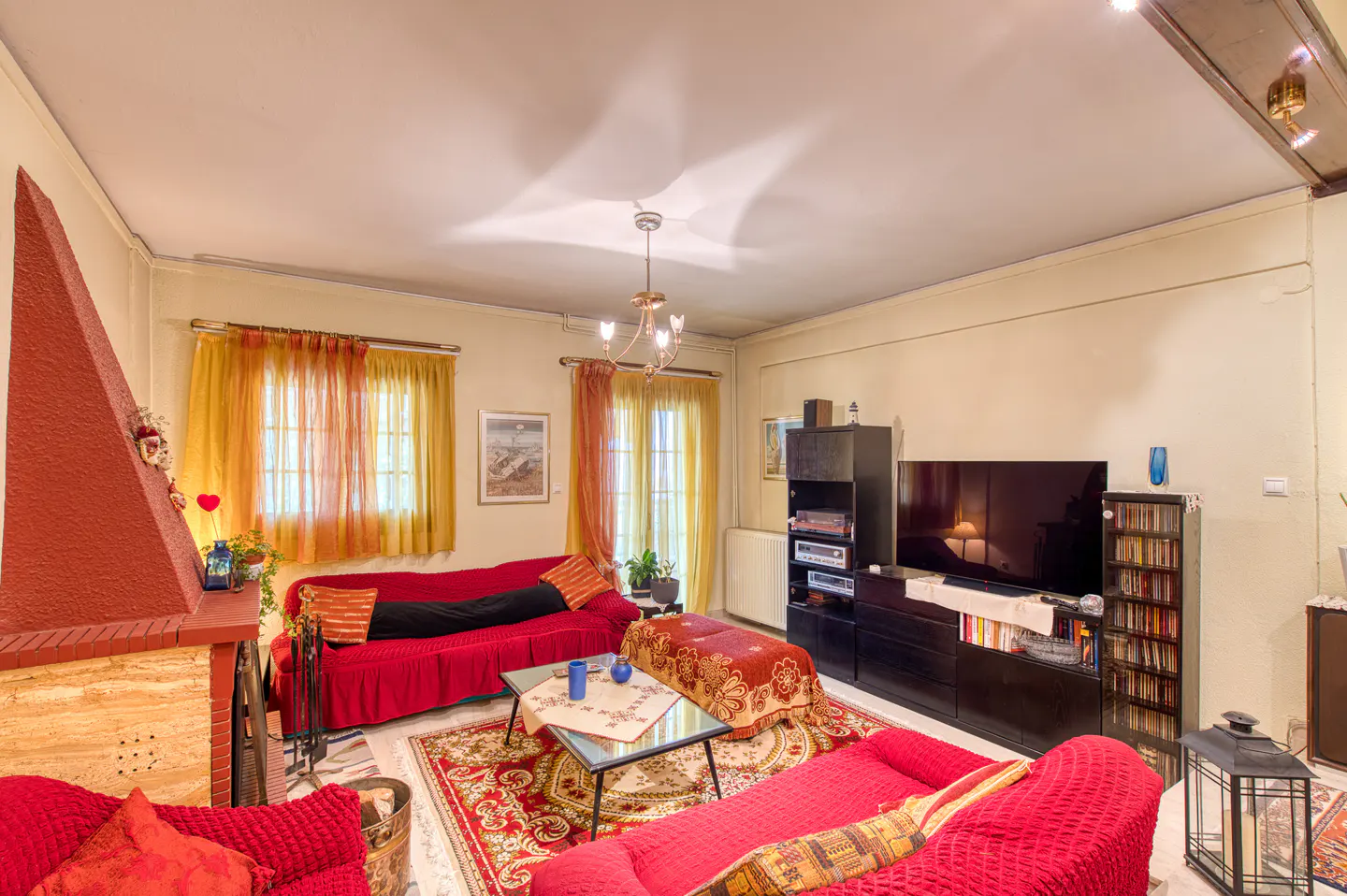 Living room with red sofas, fireplace, and TV. Yellow curtains adorn the windows. A patterned rug covers the floor.