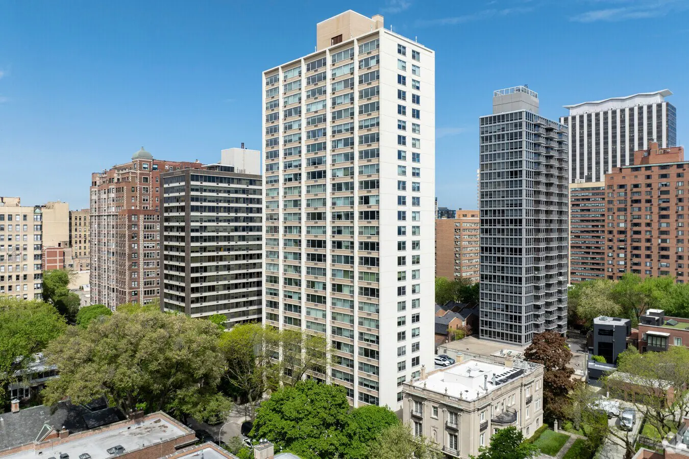 Cityscape view of tall residential buildings under a blue sky. Trees and rooftops are visible in the foreground.