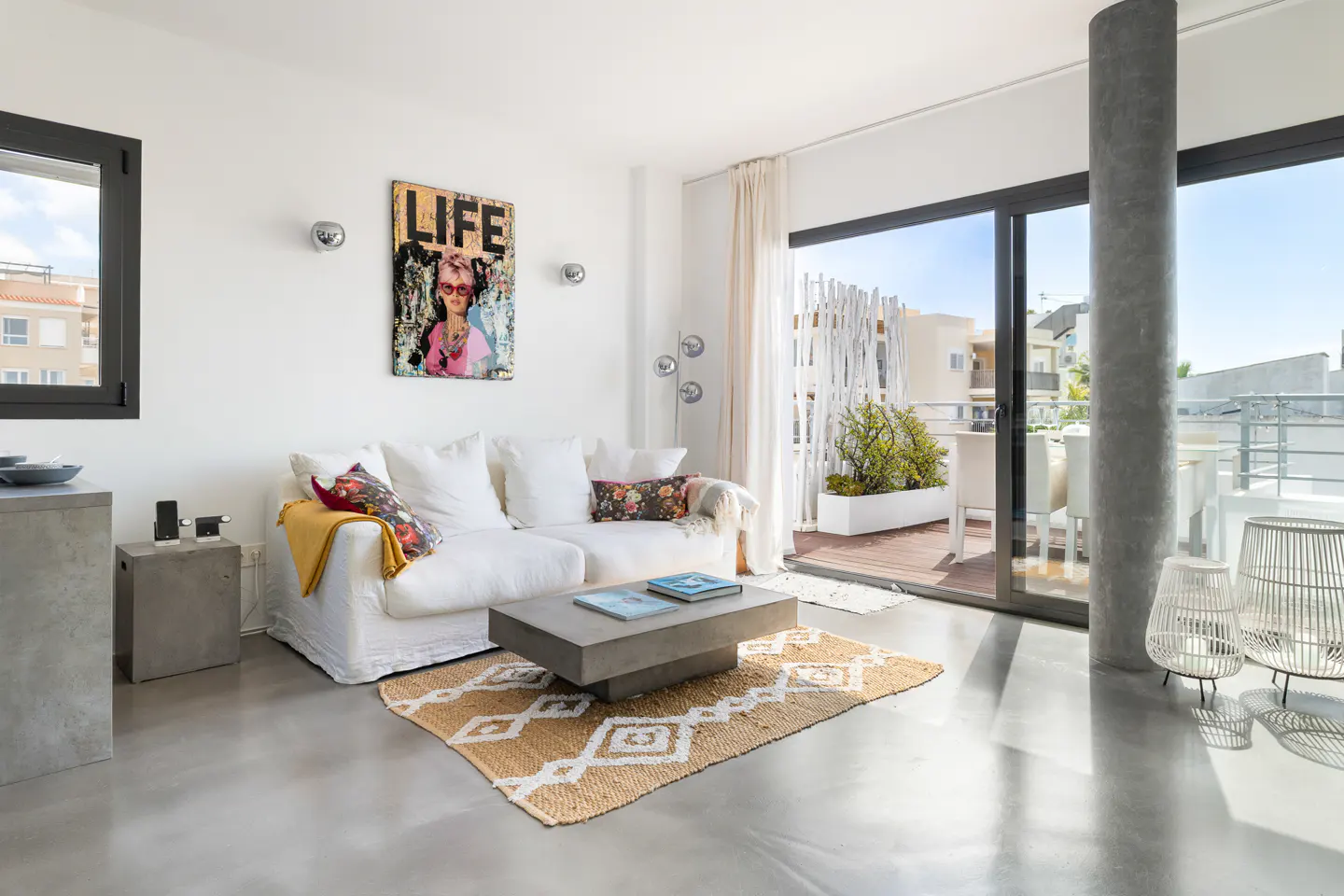 Bright living room with white sofa, concrete table on a patterned rug, and balcony access. A "LIFE" art piece hangs on the wall.