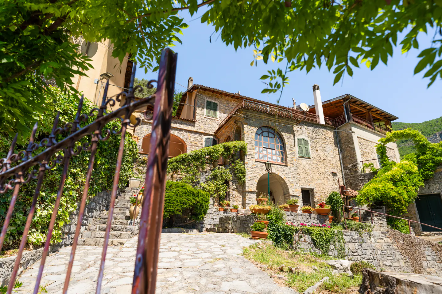 A rustic stone house with an arched entrance and stone path, viewed through a metal gate under bright blue skies.