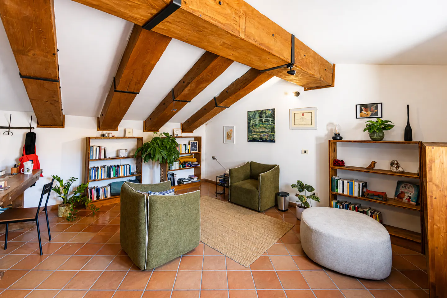 Attic living room with exposed wood beams, terracotta tile floor, green armchairs, and bookshelves.