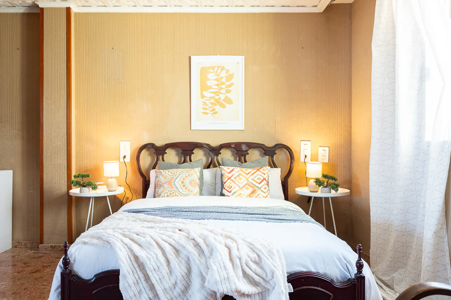 Bedroom with a dark wood bed frame, white bedding, patterned pillows, and white side tables with lamps. A botanical print hangs above the bed.