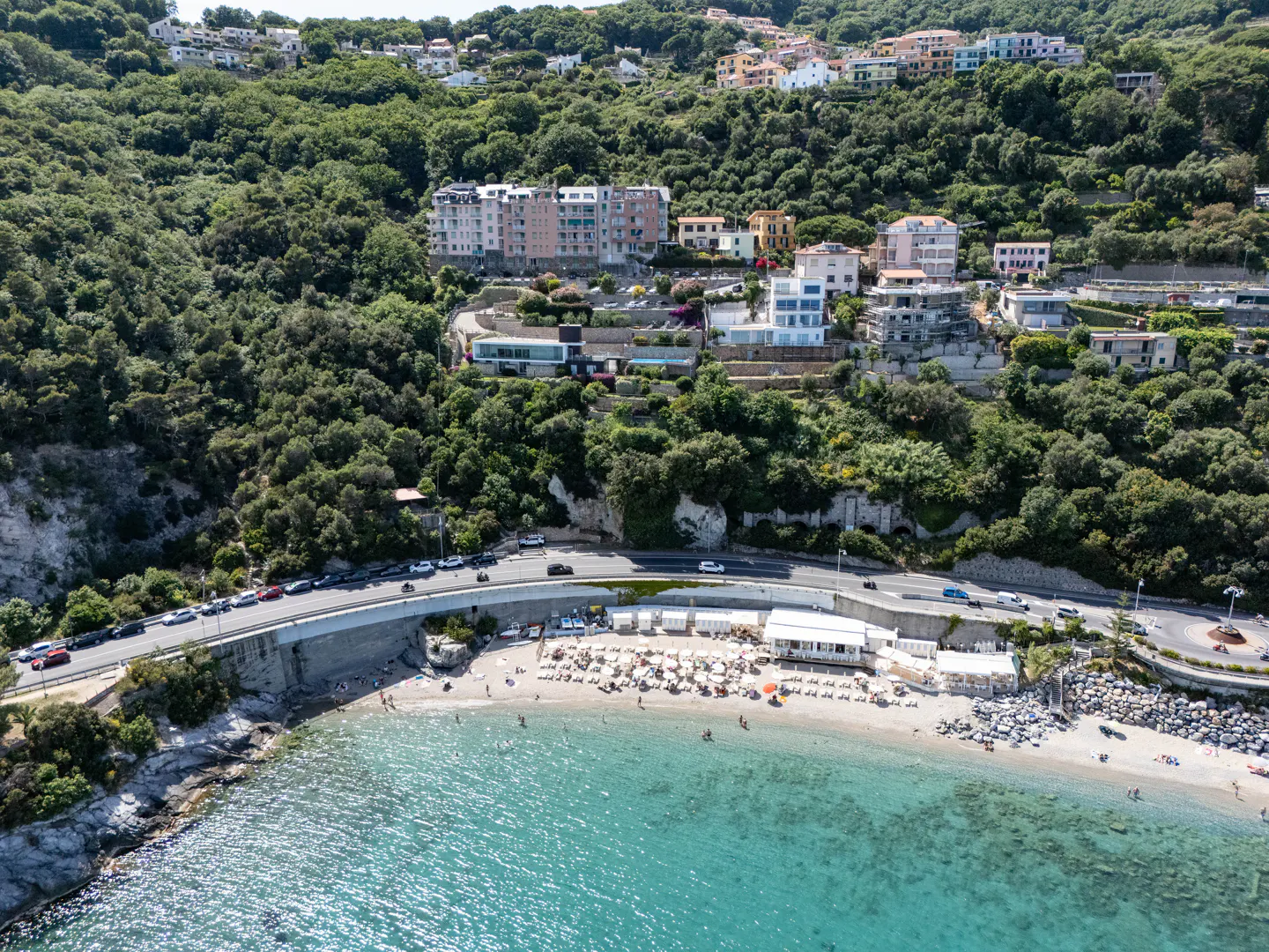 Aerial view of a beach with turquoise water, a road with cars, and buildings on a green hillside.