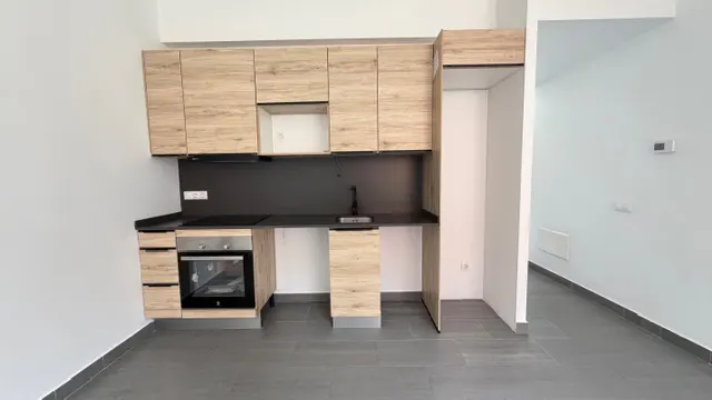 A compact kitchen with light wood cabinets, black countertops, and gray tile flooring in a white-walled room.