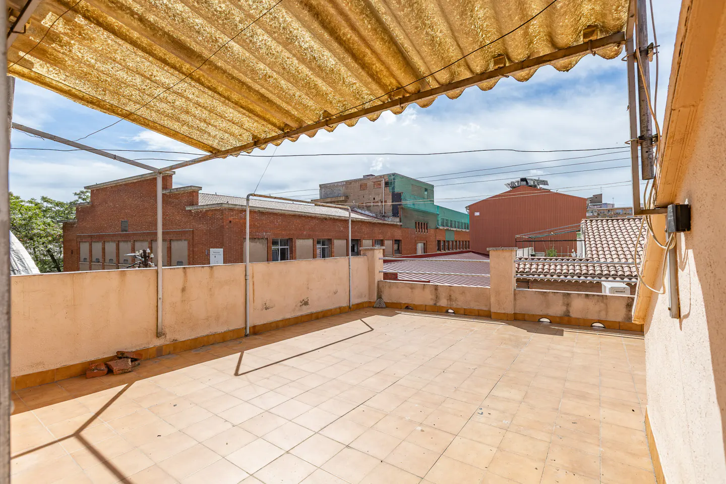 A rooftop terrace with beige tile flooring and a corrugated awning, overlooking brick buildings under a cloudy sky.